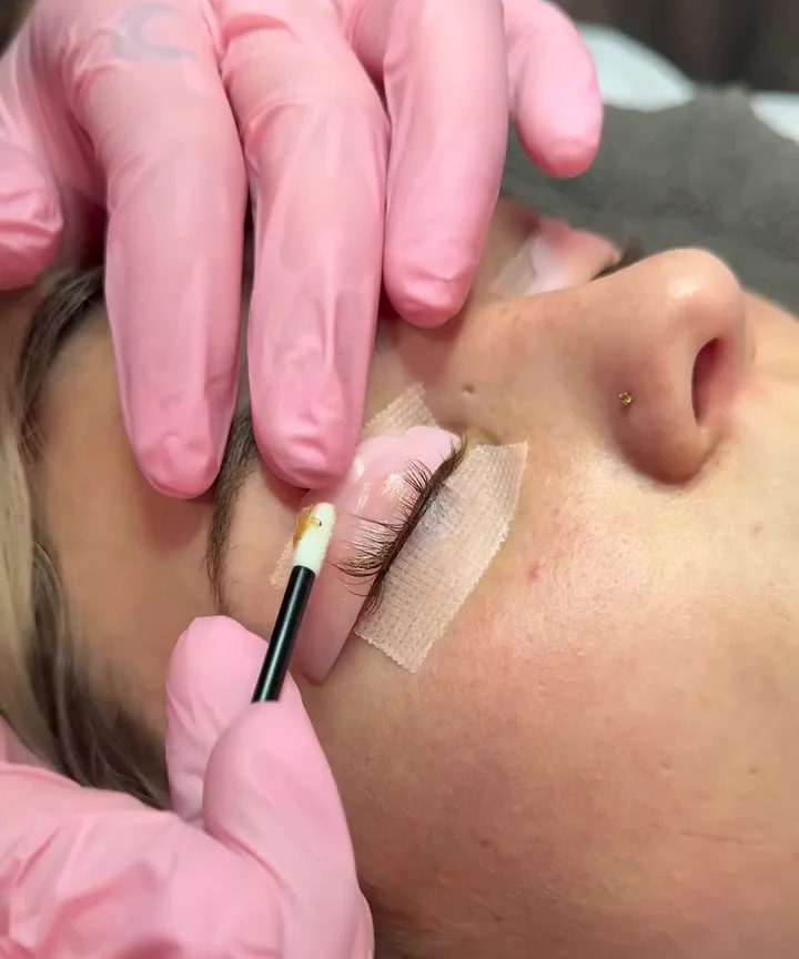 Pink gloved hands brushing lash lift solution onto a client's lashes